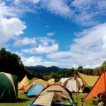 天気に恵まれた日に屋外でのキャンプイベントに大勢の方が参加されていることがイメージできるテントがたくさん張られた広大で緑豊かな草原の広場の様子